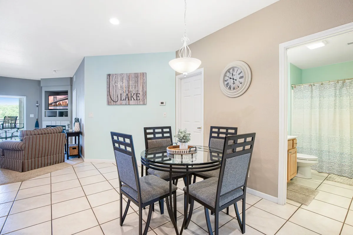 A dining area with a family-friendly table and chairs, lake-themed wall art, a wall clock, and a sofa in the background, suggesting a welcoming, communal atmosphere ideal for dining, conversation, and planning outings.