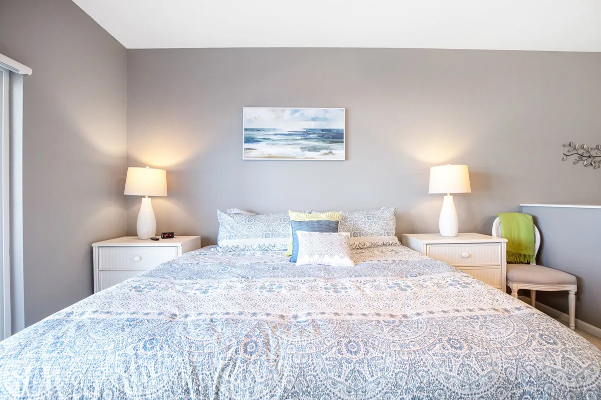 This image showcases a spacious queen bedroom with a large bed dressed in soft linens, artistic décor on the walls, and bedside tables with lamps. Natural light fills the tranquil space, enhancing the relaxing atmosphere ideal for restful sleep or reading.