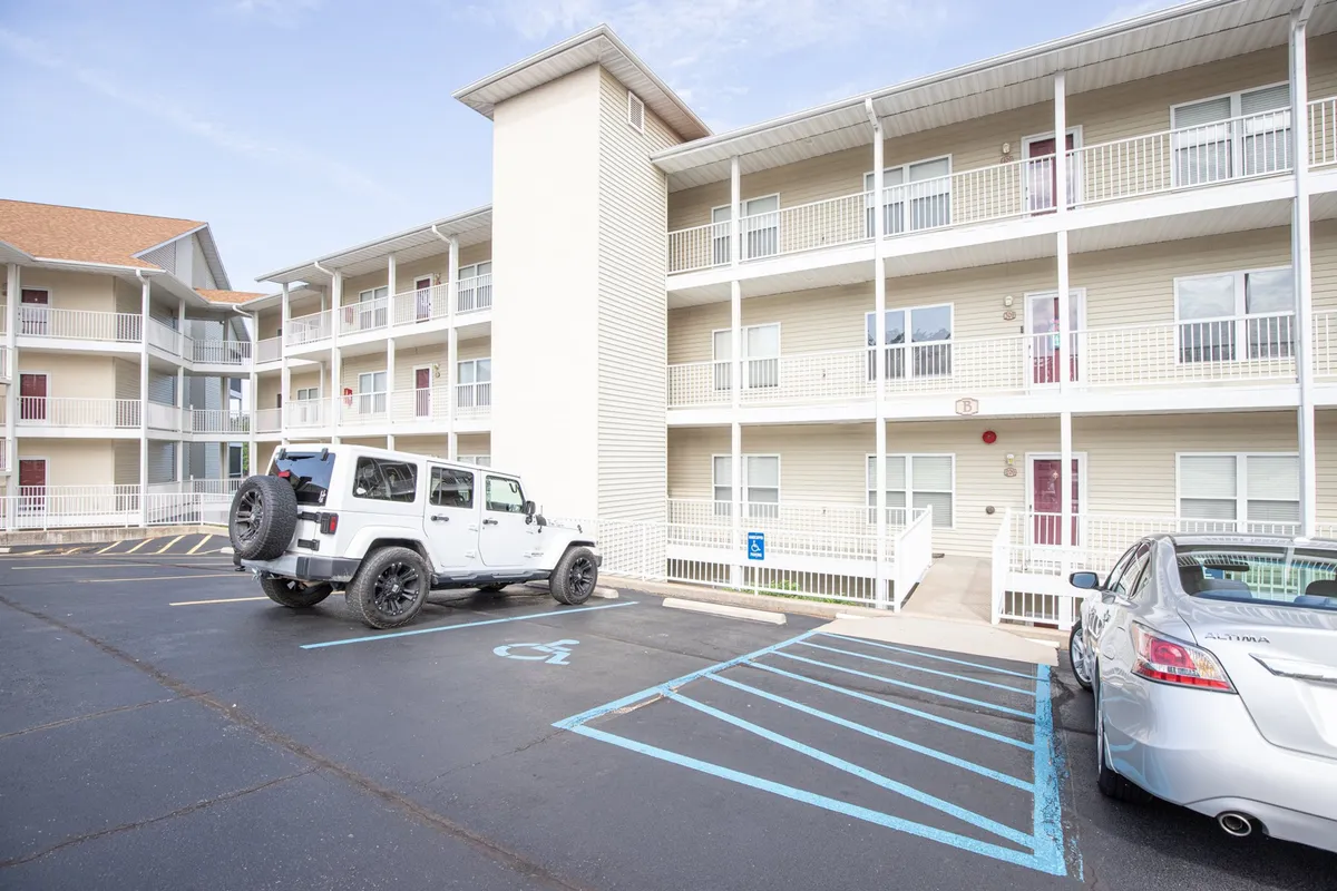 An exterior view featuring a white Jeep parked in front of a light-colored condo building with blue parking lines clearly marked. The entrance to the building is visible, suggesting convenient access for guests with mobility needs. The relaxed atmosphere hints at ease of arrival and the welcoming vibe of the property.