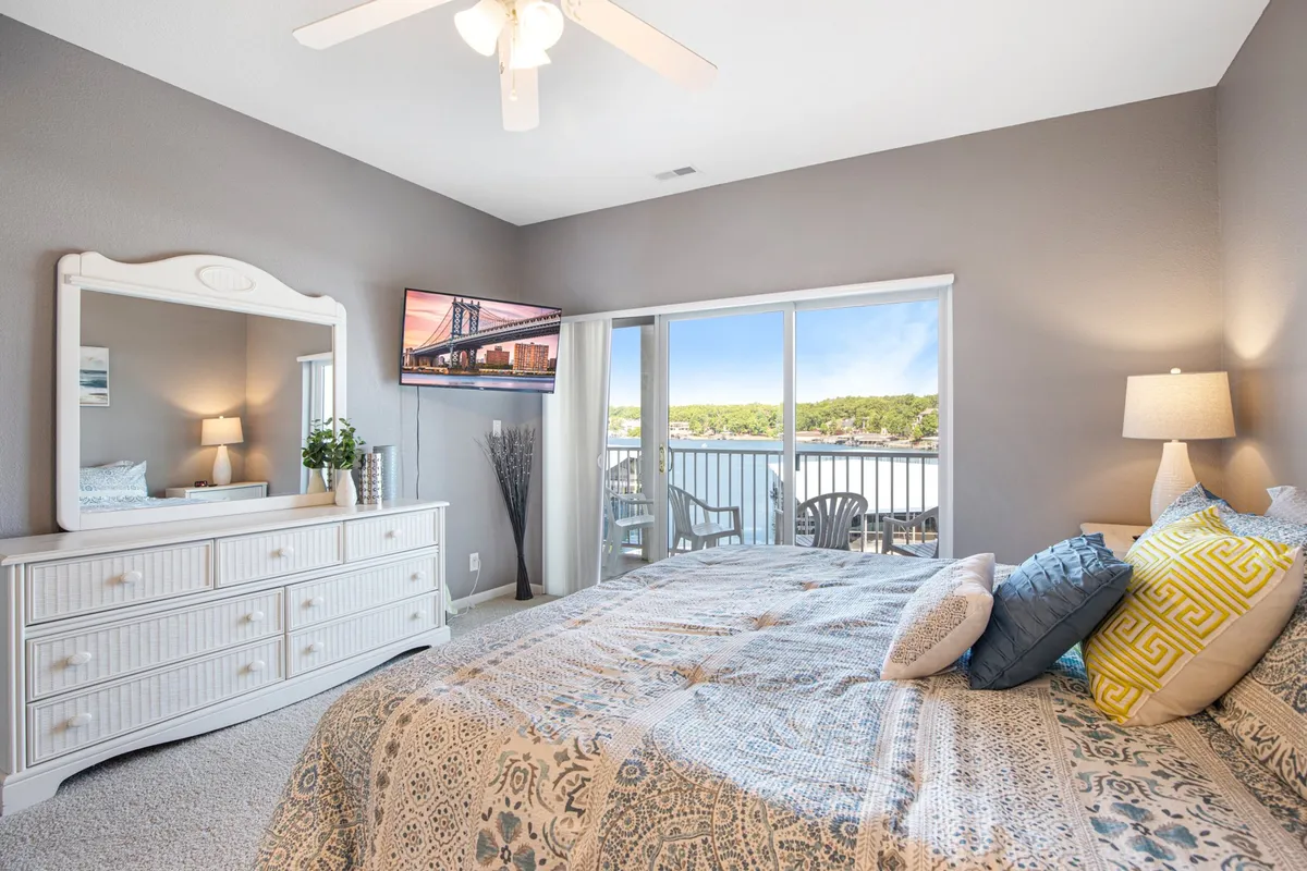 A queen bedroom featuring a bed with decorative pillows, a dresser topped with a mirror, a Smart TV across from the bed, and a sliding glass door that opens to a balcony. The room is bathed in gentle, serene light, emphasizing comfort and privacy.