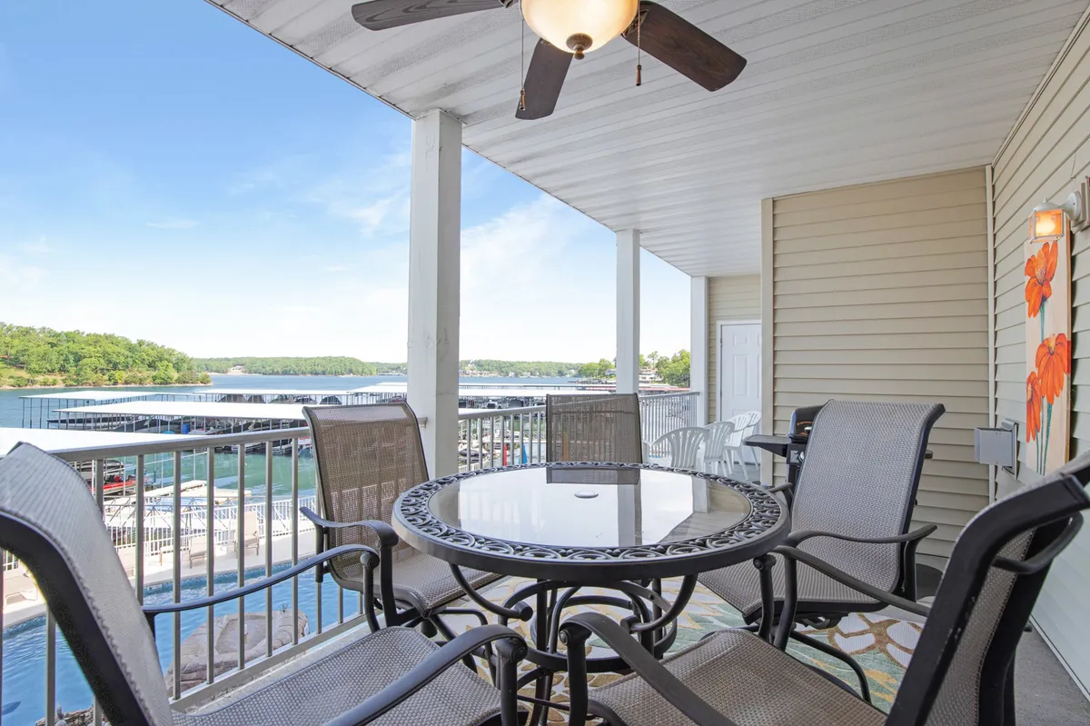The image showcases a spacious outdoor dining area on the condo's patio, complete with a ceiling fan overhead for comfortable lounging. The table is perfectly positioned to take in breathtaking, unobstructed views of the lake just beyond, with hints of sunset colors reflecting on the water, setting a tranquil, inviting mood. The scene invites guests to imagine enjoying meals, morning coffee, or evening drinks while overlooking the serene lakeside setting.